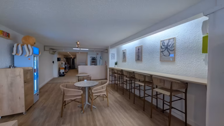 Salle avec long comptoir à tabourets, petites tables et fournil visible au fond