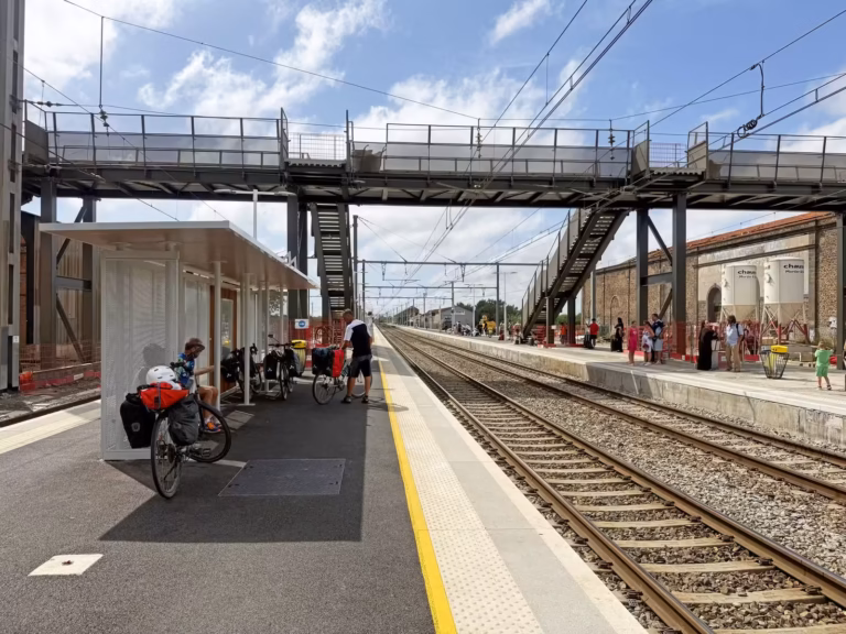 Quai de la gare d'Agde avec un abri où des cyclistes attendent, sous la passerelle, chantier en arrière-plan