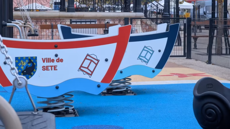 Deux jeux à ressort en forme de barques, l'une bordée de rouge, l'autre de bleu marine, marquées Ville de Sète, sur le sol bleu de l'aire de jeux