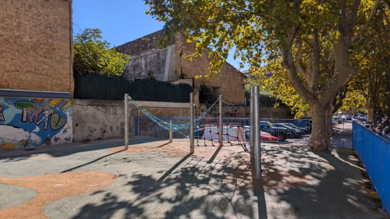 Aire de jeux clôturée devant un mur peint, sous les platanes de Poussan